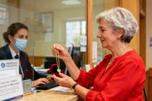 Femme âgée en blouse rouge tenant un collier en or devant le comptoir du crédit municipal de Carcassonne
