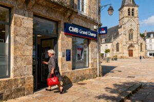 Femme de 60 ans entrant dans une agence bancaire CMB avec un sac à main rouge, façade en pierre bretonne en arrière-plan